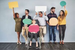 Eine diverse Gruppe von Menschen, einschließlich einer Person im Rollstuhl, hält bunte Sprechblasen hoch und symbolisiert Inklusion und Gleichberechtigung.