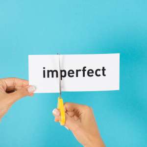 Eine Frau hat einen Zettel in der Hand mit der Aufschrift "imperfect". Diesen versucht sie gerade mit der Schere zu zerschneiden um daraus ein "perfect" zu machen.