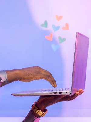 Eine Person benutzt ein lilafarbenes Laptop, auf dessen Bildschirm kleine herzförmige Symbole in verschiedenen Farben schweben, die die Idee von Online-Romantik symbolisieren.