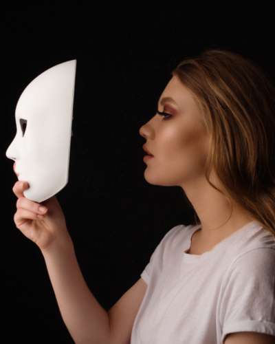 Eine Frau hält eine weiße Maske vor ihr Gesicht, die eine Trennung zwischen ihrer wahren Identität und einer falschen Identität symbolisiert.
