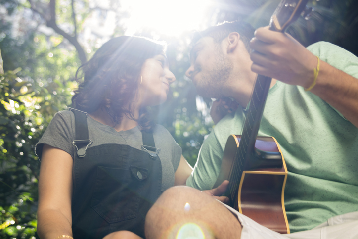 Junges Paar, das sich während der Kennenlernphase in einem sonnigen Park küsst, der Mann spielt Gitarre.