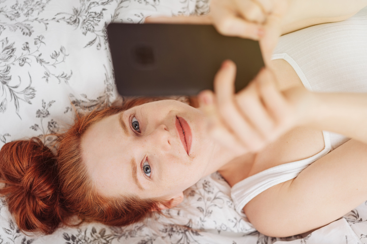 Frau mit ungeschminktem Gesicht liegt auf dem Bett und schaut auf ihr Smartphone.