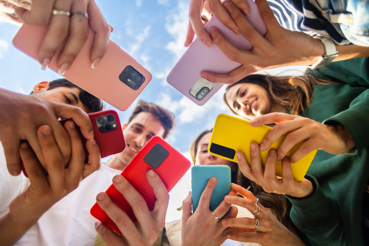 Gruppe junger Leute mit Smartphones in verschiedenen Farben, die gemeinsam lachen und etwas auf ihren Bildschirmen betrachten – repräsentiert die Testphase der Diner Date App.