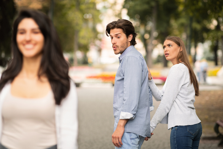 in Mann schaut einer anderen Frau hinterher, während seine Partnerin ihn verärgert zurückhält.