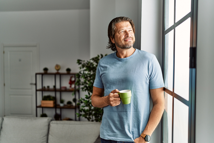 Glücklicher Single Mann genießt Kaffee am Fenster und blickt nachdenklich in die Ferne.