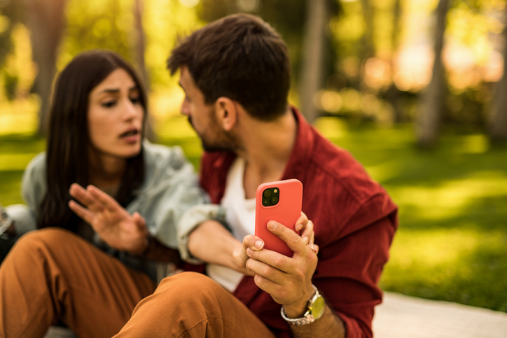 Ein Paar diskutiert im Park, während der Mann ein Smartphone hält, das Misstrauen und Eifersucht symbolisiert.