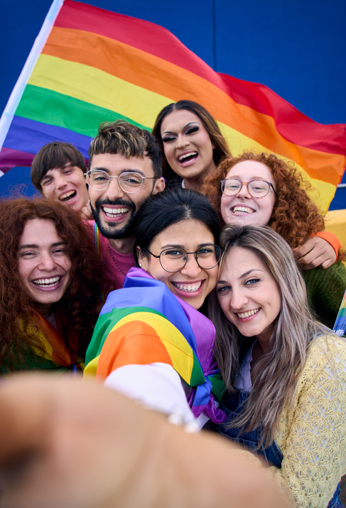 Gruppe junger LGBTQ+-Personen mit Regenbogenflagge, die gemeinsam in die Kamera lächeln. Symbolisiert Vielfalt, Gemeinschaft und politische Diskussionen.