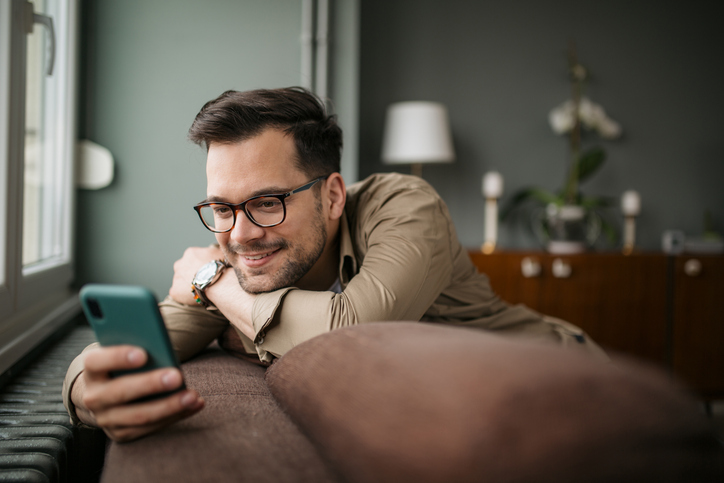 Mann mit Brille liegt entspannt auf dem Sofa und lächelt beim Blick auf sein Smartphone.