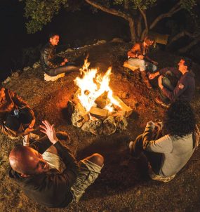 Sechs junge Leute sitzen im Kreis um ein Lagerfeuer und unterhalten sich gut gelaunt.