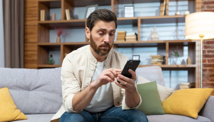 Ein nachdenklicher Mann sitzt auf einem Sofa und schaut auf sein Smartphone, während er eine Dating-App benutzt.
